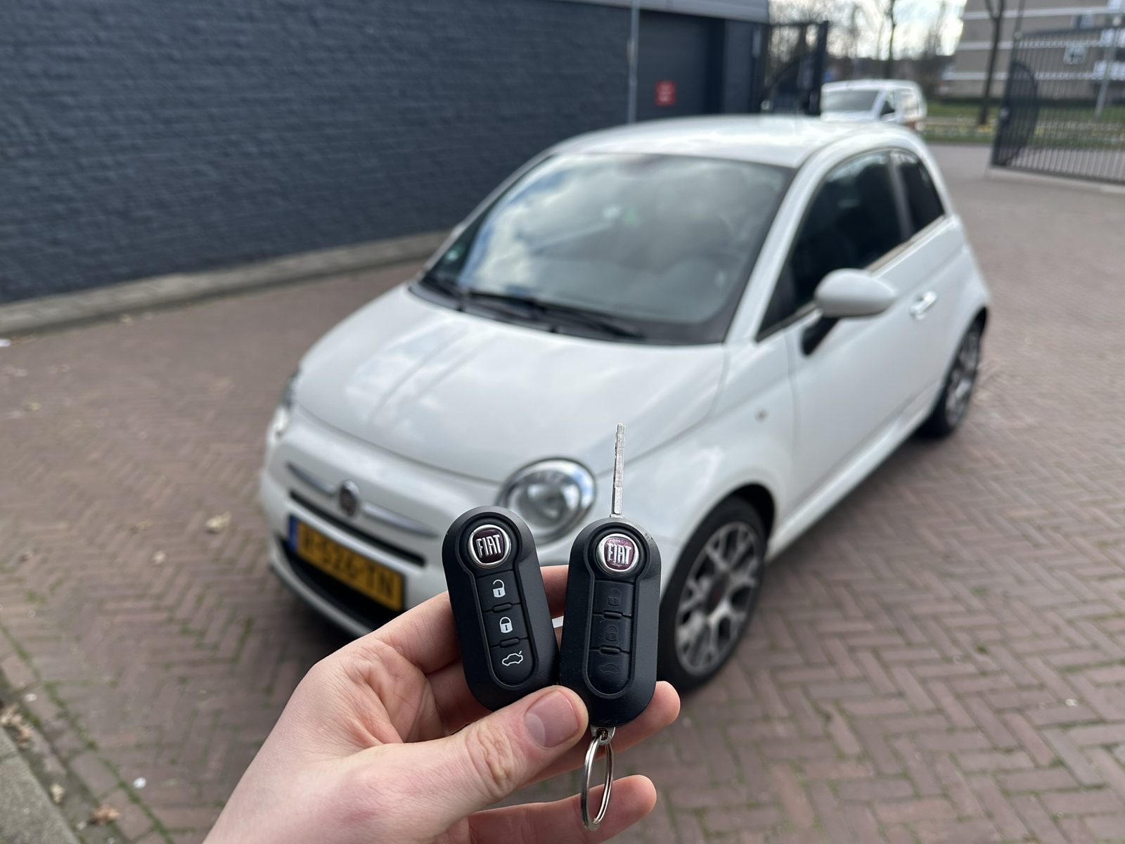 Twee Fiat-klapsleutels in de hand, met de witte Fiat 500 op de achtergrond